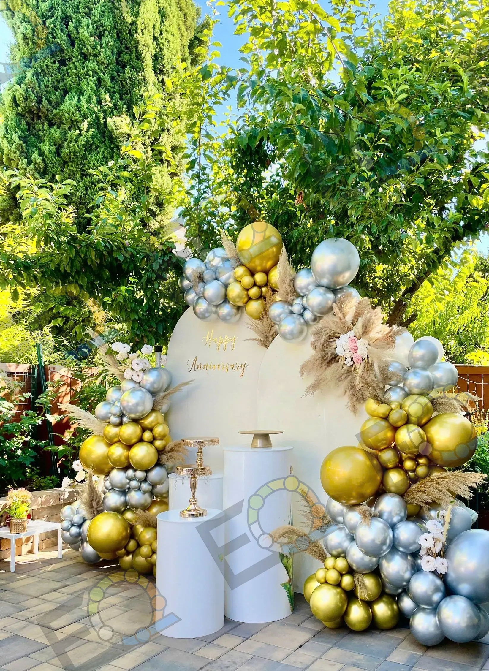 A gold and silver balloon arch around a white anniversary backdrop in a garden